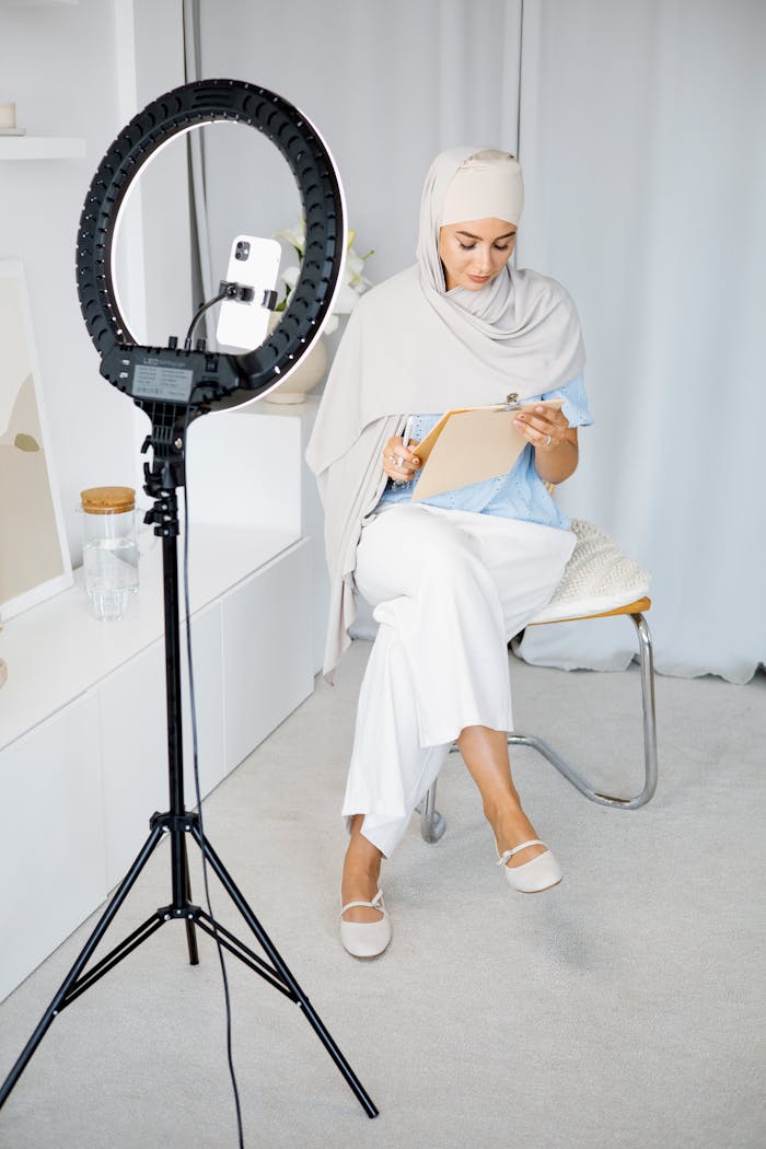 Woman in hijab sitting with clipboard, recording content with ring light.