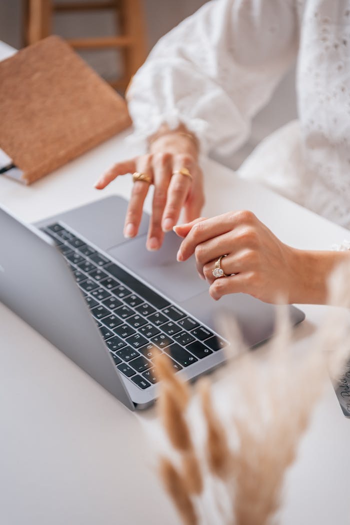 Close-up of a woman's hands typing on a laptop, adorned with rings, in a cozy indoor setting.